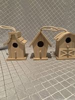Front view of three unpainted mini wooden birdhouses on a patterned background showing hanging ropes and various roof styles.