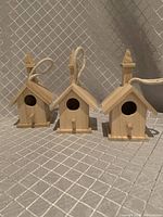Three identical unpainted wooden mini birdhouses lined up side by side showing front and side features with rope loops for hanging.