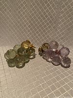 Two clusters of acrylic grapes on copper wire, one light green and one light purple. Both have faceted spherical grapes and gold-colored leaves, against a textured background.