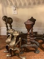 Three sets of brass andirons in varying sizes placed on wooden floor near electrical outlet showing condition and size comparison.