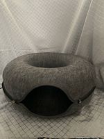 Gray felt cat tunnel bed with circular donut shape, zipper partially open showing oval entrance, photographed against a light grid backdrop.