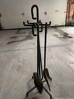 Full view of fireplace tool set with stand including poker, brush, shovel, and another tool, standing in garage on concrete floor.