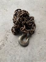 Coiled heavy-duty metal chain with a large metal G-70 hook attached, showing surface rust on the chain links and hook.