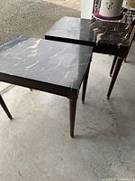 Pair of square side/end tables with thick black marble tops showing beige veining, standing on tapered wooden legs with brass feet and accents.