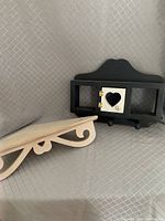 Black mini cupboard with white heart cutout door and two pegs below, resting on a light wood decorative corner shelf with cutout brackets.