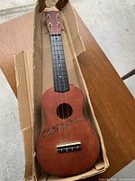 Full view of Duke Kahanamoku brown wooden ukulele packed in its original cardboard box on a wooden table.
