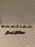 Photo of Pontiac and Grand LeMans car badges on a quilted fabric background, showing front side with logos.