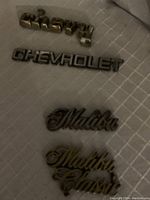Four different vintage car badges: Chevy, Chevrolet, Malibu, and Malibu Classic, displayed on fabric surface.