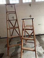 Two wooden step ladders, one 5-step and one 3-step, standing in a garage against closed garage doors.
