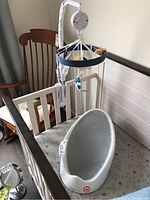 Baby bath support placed inside a crib with an attached white arm holding the baby mobile with hanging toys (cat, fish, yellow round toy).
