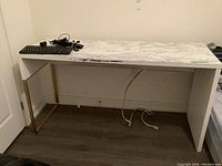 Full view of desk showing marble-pattern surface, white wood frame, gold metal frame on left side, white wood panel on right. Keyboard and cords on top for scale.