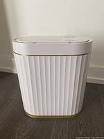 Front view of white Elpheco electronic garbage bin with vertical ridges and gold trim on a dark floor.