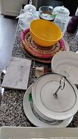 Three covered glass dishes, glass ice bucket with handle, yellow plastic bowl, faux marble bowl, cake stand, cheese slicer board, and woven tray on a granite countertop