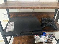 Sony PlayStation 3 slim console with two wireless controllers on a wooden shelf, visible game cases under shelf.