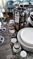 Photo of the two-level wooden spice rack with eight small glass spice bottles and one taller glass bottle, all with silver lids, arranged on a kitchen counter next to stacked plates.