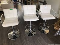 Three white faux leather adjustable stools with chrome bases, seen from front, showing their overall appearance and condition including some wear.