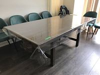 Full view of rectangular wood veneer table with clear plastic cover, surrounded by several green upholstered chairs. Table fully extended with leaf in place.