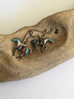 Pair of silver horse-shaped earrings with turquoise, coral, opal, and goldstone mosaic inlay on a wooden display.