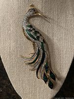 Photo of the gold tone peacock brooch displaying rhinestones and green and black enamel accents on a neutral fabric background.