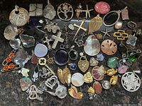 Photo showing an assortment of approximately 57 pendants laid out on a dark surface. Various designs and materials including metal, stone, glass, enamel, and shell are visible.
