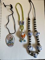 Photo showing three handpainted porcelain jewelry pieces on white background: oval pendant with woman figure, bead with elephant design on yellow cord, and blue and white bead necklace with brass inserts
