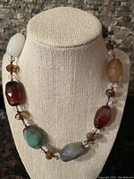 Necklace displayed on beige textured bust showing arrangement of multi-coloured stones and faceted crystal beads.