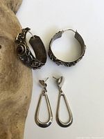Two pairs of sterling silver earrings shown including one pair of wide Brutalist hoops and one pair of oval dangle earrings.