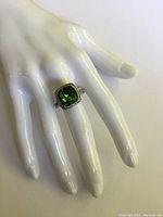 Ring worn on white mannequin hand to show scale and fit, highlighting the green center stone and filigree band.