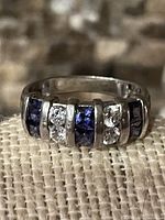 Close-up front view of ring showing 6 square-cut purple tanzanite stones alternating with 6 clear stones, all channel set in a silver band.