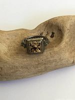 Front and side view of ring showing square top with raised flower design, gold-tone accents, and reddish-brown stone.