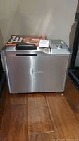 Side/front view of the Breville bread maker showing the stainless steel exterior and the recipe book on top