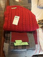 Photo shows folded red knitted throw blanket on top of a folded patchwork quilt with two pillow shams underneath.