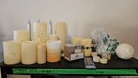 Variety of cream-colored battery operated pillar candles, some with carved floral or snowflake patterns, small battery operated tealights, packs of candle batteries, and plastic wrapping materials arranged on a table.