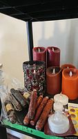 Photo showing multiple large cylindrical battery operated LED pillar candles in red, brown, and orange, along with wrapped brown wax sticks and a metal candle holder.