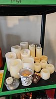 Overhead view of various battery operated and handmade pillar candles along with tealight battery operated candles and button cell batteries on a green shelf.
