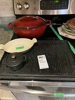 Full view of red cast iron casserole dish, small black Staub pot, ceramic dish, and grill tray on stove