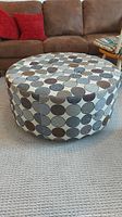View of the round upholstered ottoman showing all-around patterned fabric in circular designs, placed on carpet in front of brown sofa.
