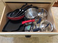 View of drawer filled with pots, pans including three frying pans (black and red), a square grill pan with red handle, stainless steel steamer insert, and glass lids with blue handles.