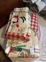 Photo showing folded Christmas quilt with snowman appliqué and red plaid border, along with holiday motifs in background, close-up view