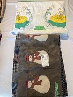 Two quilts folded and displayed, one predominantly white with yellow flower embroidery, another dark patchwork with snowmen appliques, both hand quilted and machine pieced.