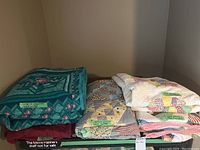 Image showing a collection of folded quilts stacked in two piles with labels noting quilting method (hand quilted, machine pieced).