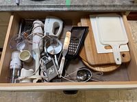Overview of drawer with all kitchen items including hand mixer, grater, cutting boards, measuring cups, icing spreader, and thermometer.