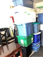 Stack of nine plastic storage totes in various colors with lids and one open black bin at the bottom, positioned near black chairs and books.