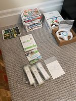 Image showing stack of Wii game cases, several titles readable include CSI, Wii Fit, Wii Sports Resort, Mario Party, others. Four Wii remotes and Wii Wheel in box beside games.