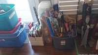 Photo showing a large storage tote filled with rolls of wrapping paper and holiday-themed packaging materials, cardboard box with wrapping paper roll tubes, and stacked plastic storage bins.