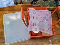 Orange plastic picnic box opened showing nested Tupperware containers and translucent plastic mat with gingerbread man graphic.