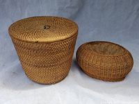 Photo showing two woven baskets: one large cylindrical covered basket with loop handle on lid and one smaller oval open coiled basket