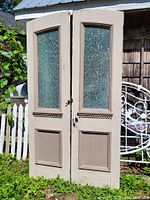 Pair of beige pine double doors displayed outdoors showing the full front view with textured glass panes, decorative wood panels, and hinges.