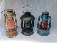 Full view of three vintage barn lanterns showing varying colors and conditions including rust, paint wear, and different globe colors