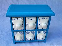 Front view of small blue wooden chest with six white ceramic drawers featuring blue decorative designs and round knobs, each drawer contains a molded scented candle inside.